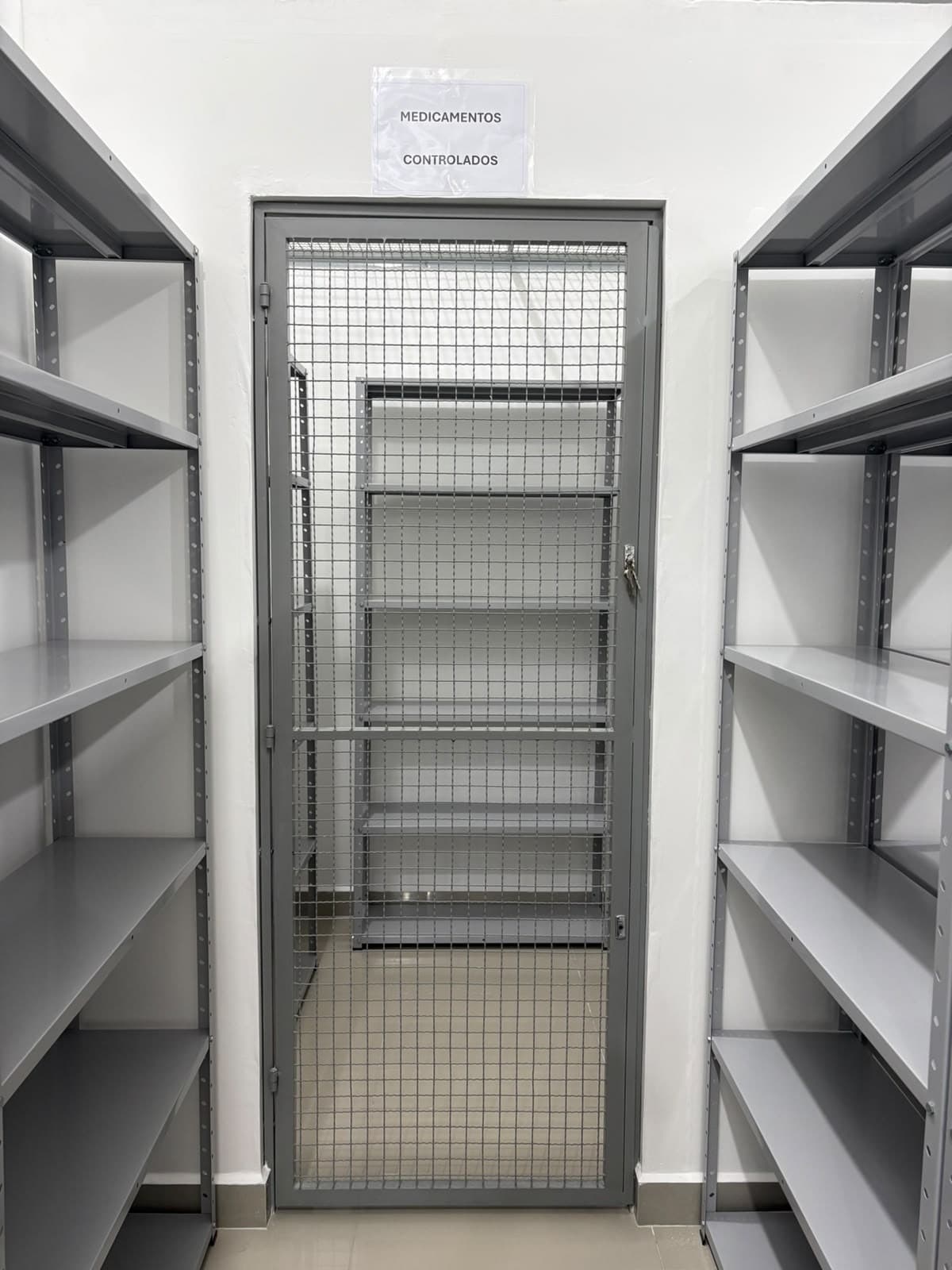 Área segregada de medicamentos controlados, com porta gradeada e estantes metálicas.